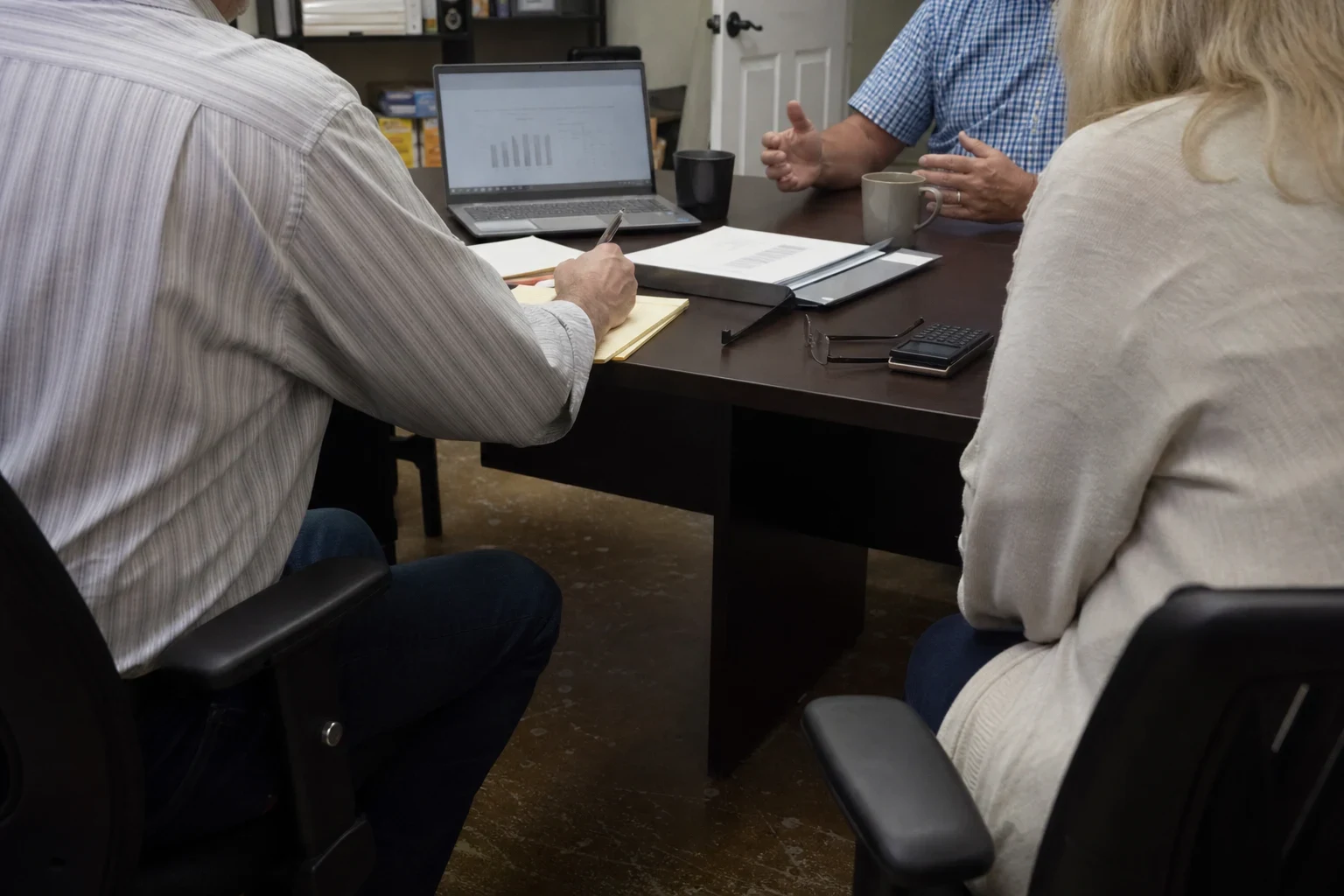 Man in a striped shirt takes notes on a legal pad at a dark conference table as a man in a blue plaid shirt talks and a woman listens, laptop showing a bar chart