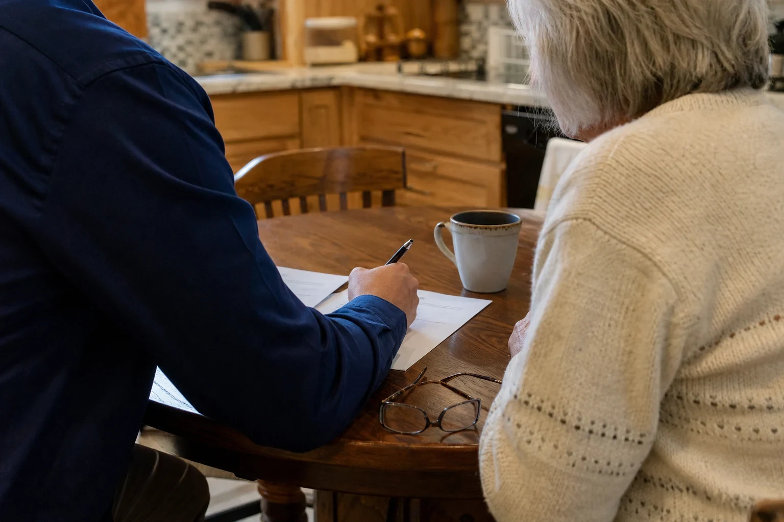 Agent in a navy shirt writes on a form at a wooden kitchen table beside an older woman in a cream sweater, with a coffee mug and reading glasses nearby