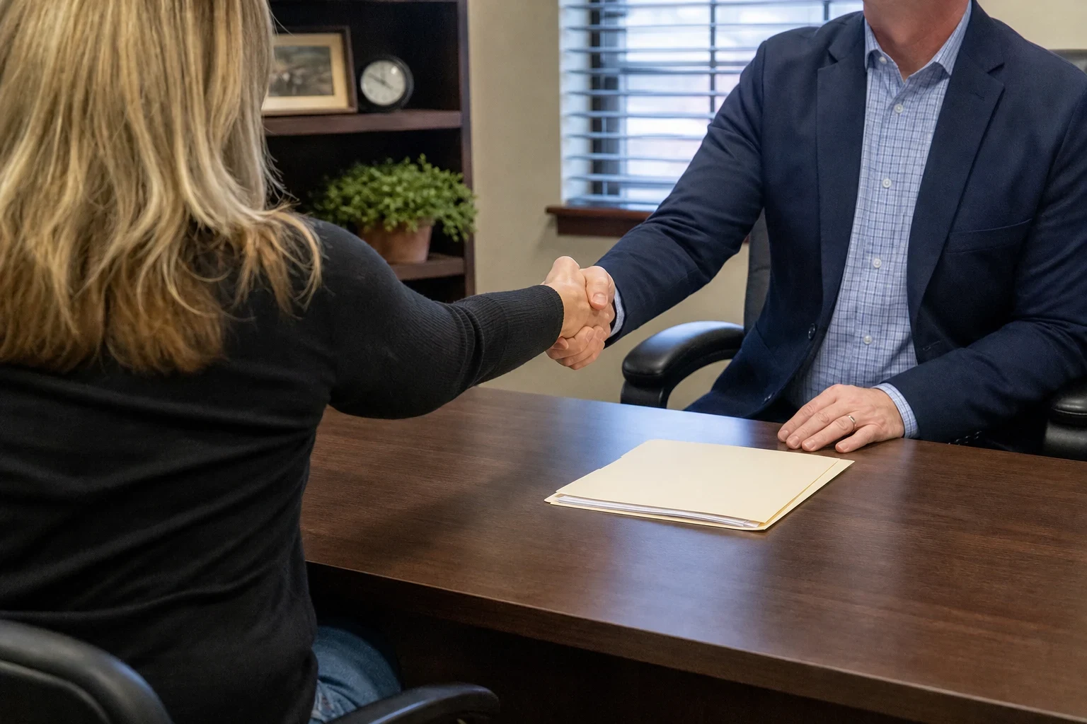 A woman and a man in a navy blazer shake hands across a dark wood office desk with a manila folder, bookshelf with plant and clock and a window with blinds behind