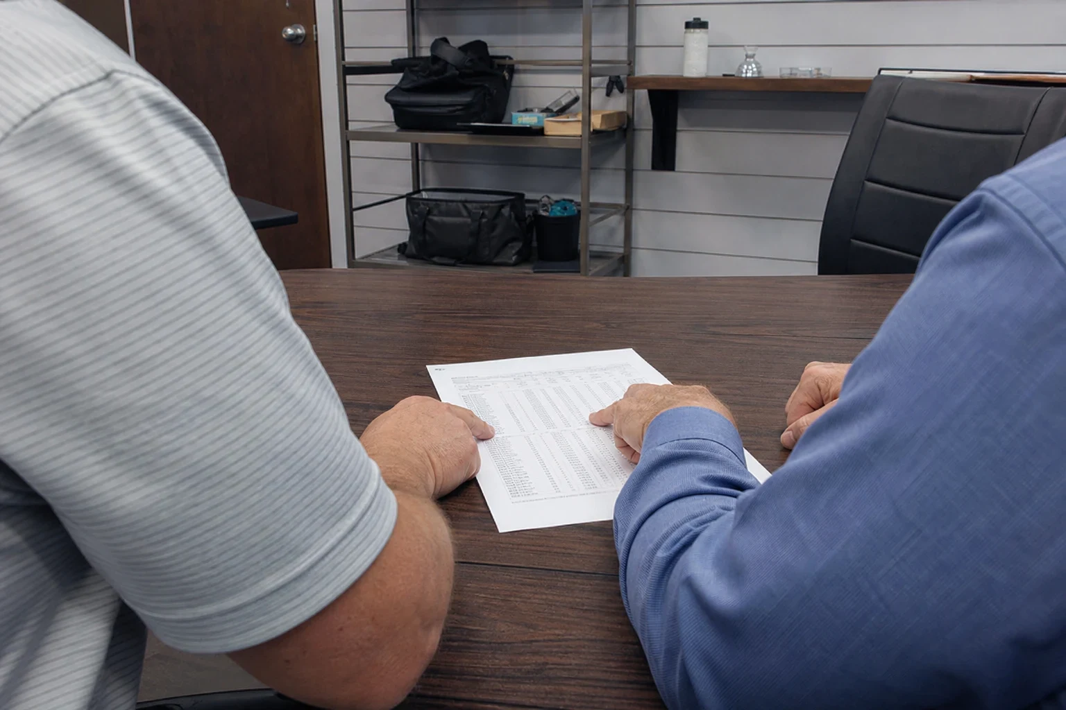 Two men seated at a dark wood conference table reviewing a printed table of figures, with metal shelving, bags and an office chair in the background.
